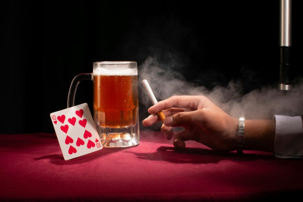 pexels-photo-36673068-36673068 A hand holding a cigarette beside a beer and playing card on a red tablecloth.