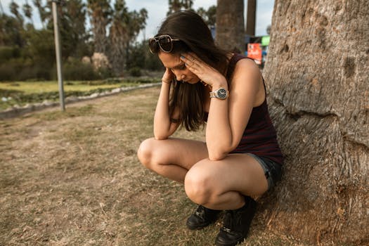pexels-photo-5542968-5542968 A woman kneels by a tree holding her head, expressing stress or anxiety in an outdoor park.