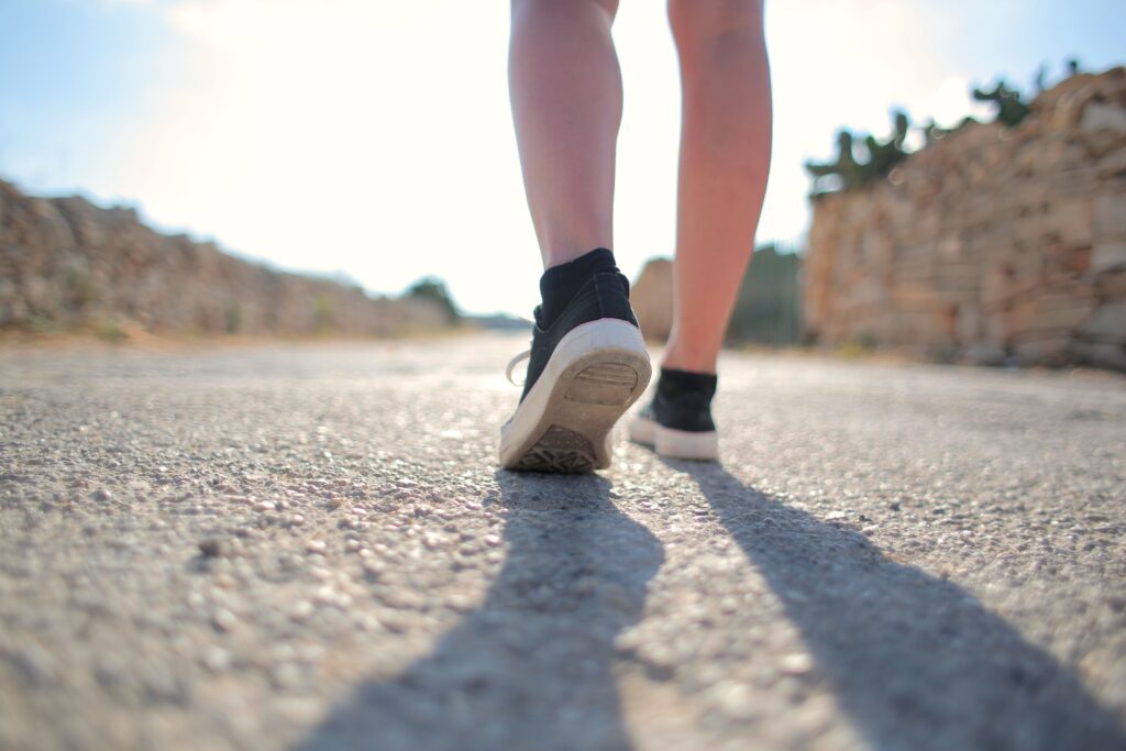 pexels-photo-3785405-3785405 A person walks on a sunny rural road wearing sneakers, casting a long shadow.
